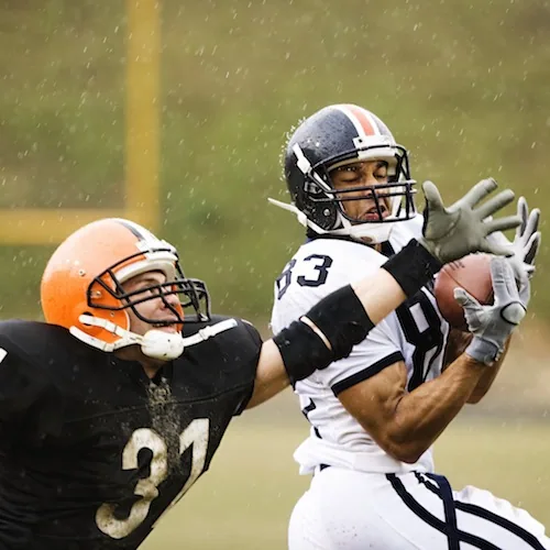 Football player in white uniform catching ball while defended by player in black uniform in rainy conditions